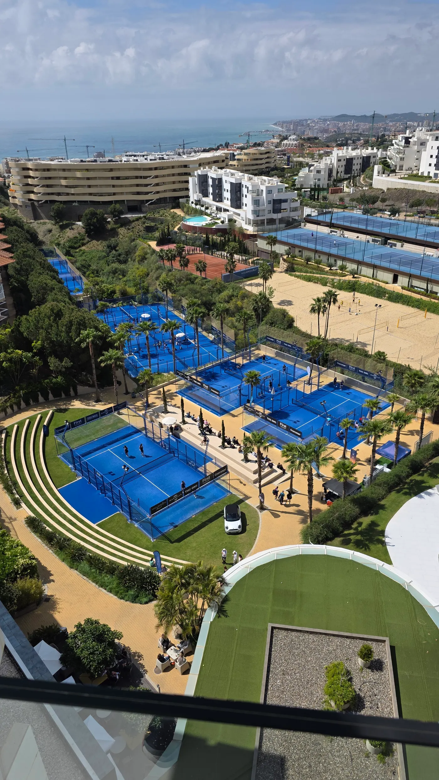 View from the El Higueron hotel rooftop in Fuengirola — multi-court padel facility with palm trees, swimming pool, and ocean beyond.